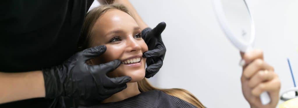 Smiling girl looks in mirror while cosmetic dentist points to her teeth.